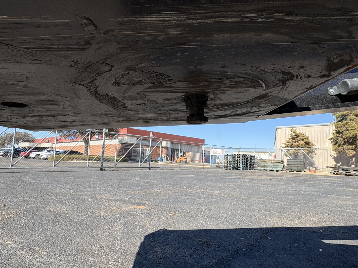 Underbody view of a Load King trailer showing the underside and hitch area with gravel lot and buildings in the background.