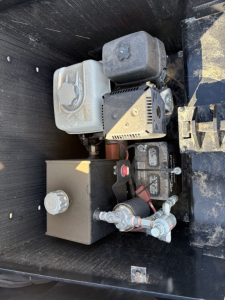 Trailer component image showing small gasoline engine with fuel tank, hoses, and hydraulic or pump assembly in a lowboy trailer toolbox compartment.