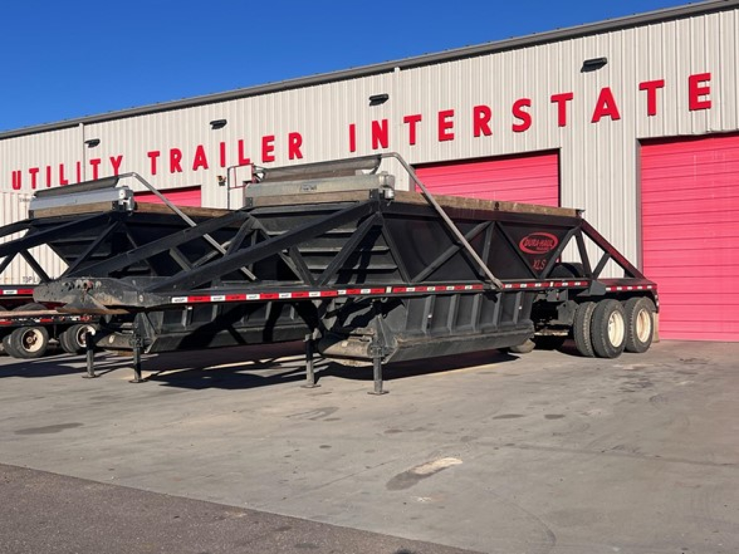 Bottom dump trailer with air ride system parked outside a facility with red garage doors and 'UTILITY TRAILER INTERSTATE' sign in the background.