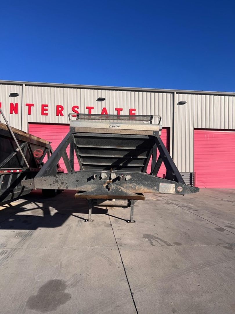 Truck trailer bottom-dump hopper with steel frame and hitch, resting on concrete in front of a building with red doors; no visible text on the equipment surface.