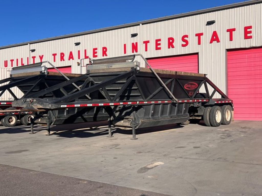 Dump trailer showing low-angle side view with black frame and tandem axles, parked in front of a red-bay building labeled 'Utility Trailer Interstate'.