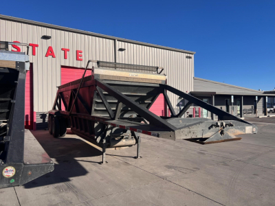Bottom-dump trailer on a concrete lot with a metal frame and ramp, in front of a beige building with red doors.