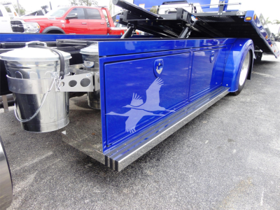 Blue Peterbilt rollback truck side with fuel tank and graphic logo showing white bird silhouette on door panel.