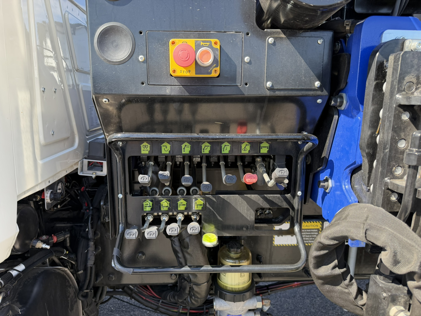 Mack GR64F truck engine bay with control switch panel showing red emergency stop, orange button, and multiple green labeled levers/valves. Battery area and hoses visible.