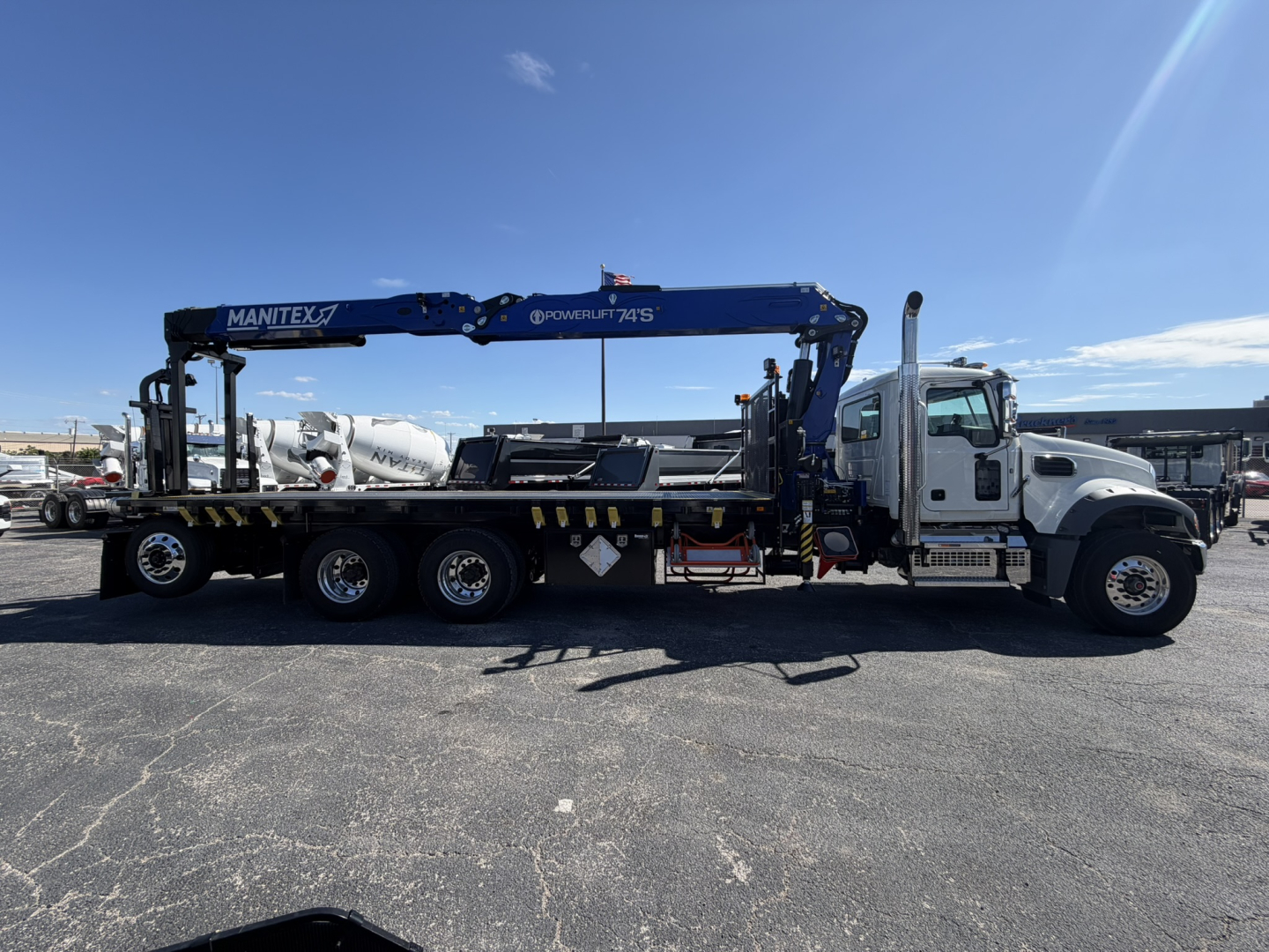 Truck chassis with a long crane/stinger, Mack GR64F, white cab, blue crane arm labeled Manthey and POWER LINES, side mirrors, stake bed on flatbed trailer.