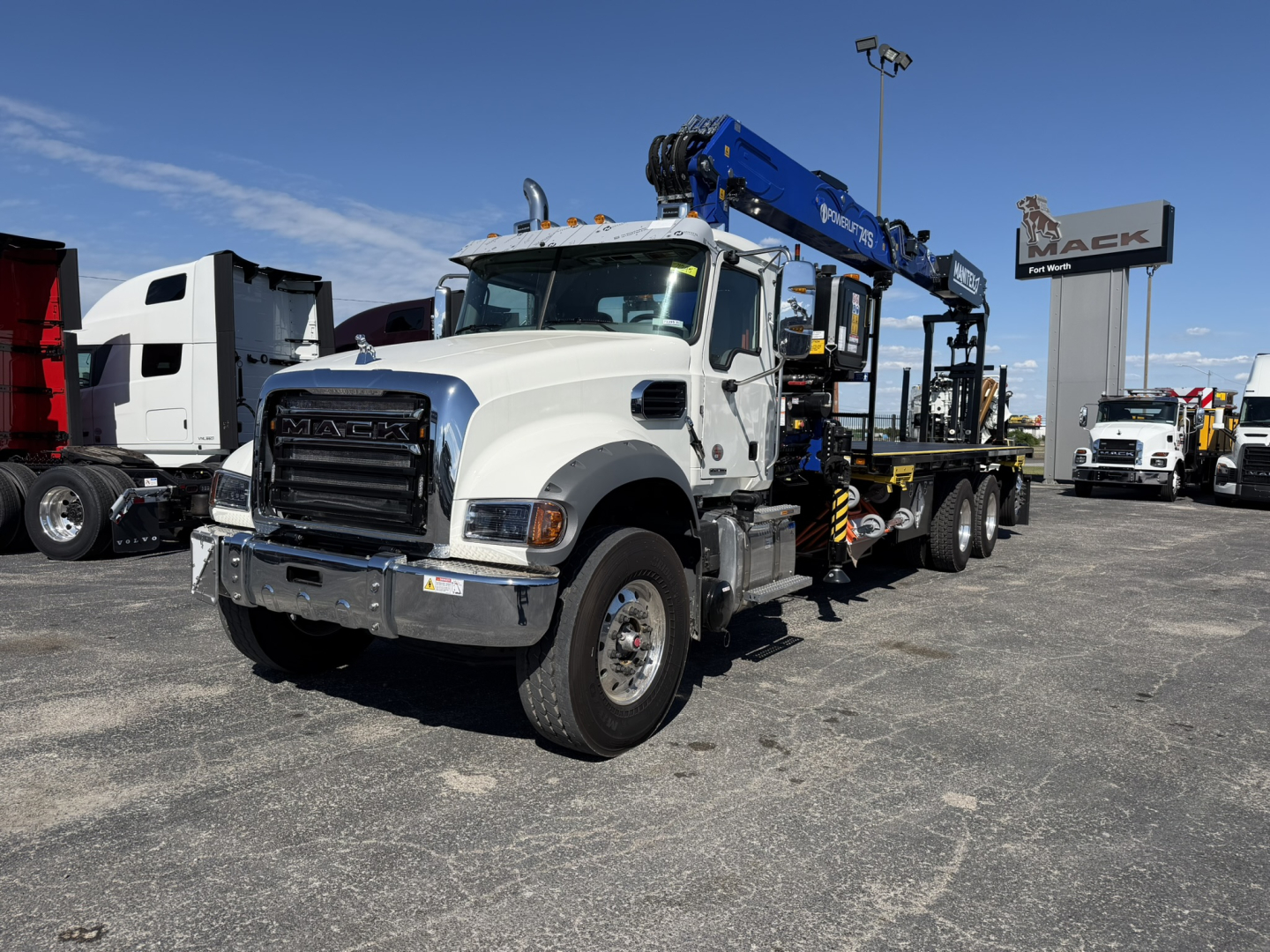 Truck cab showing Mack branding and a blue crane boom mounted on a white truck chassis; surrounding lot with other trucks and a Mack sign.