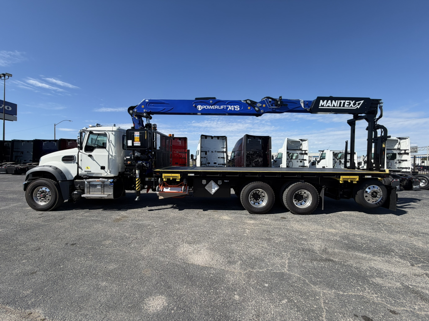 Truck-mounted crane on a Mack GR64F flatbed tow truck with a blue Manitex boom extended, side view showing cab and dual rear axles.