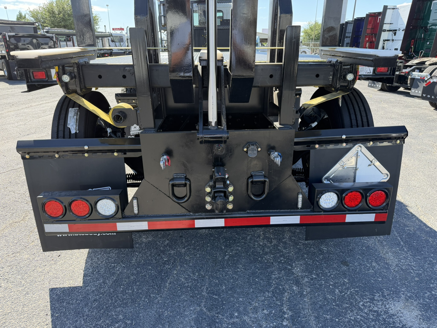 Rear view of a Mack GR64F truck chassis with hydraulic lift forks, safety chains, and reflective tape; multiple lights and triangular reflective sign on bumper.