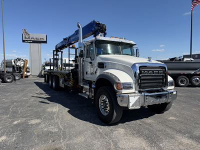 Mack GR64F heavy tow truck with a blue crane in an outdoor lot. Front grille displays MACK. Visible text includes Mack Powers for Hire on sign in background, and truck features like chrome bumper and white cab.