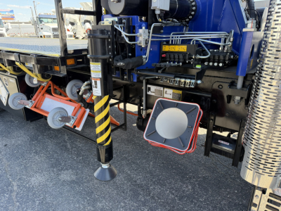 Crane truck rear with stabilizer leg and control components; visible labels include safety stripes on leg and circular white horn-like component, along with hydraulic lines and gauges.