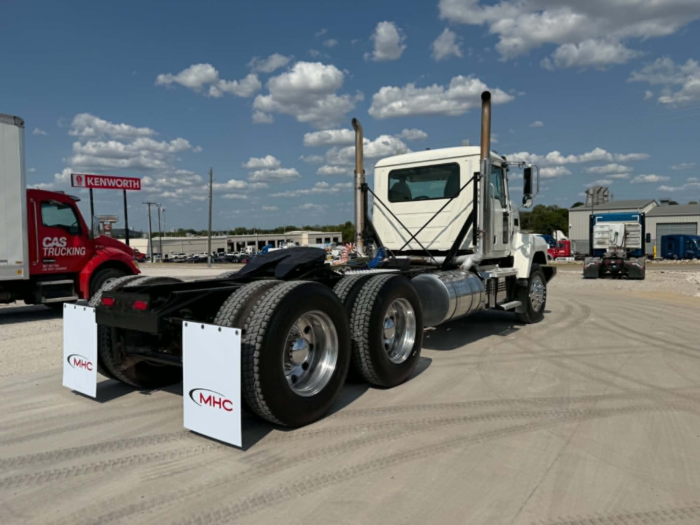Mack CHU613 daycab tractor rear view showing dual rear axles and exhaust stacks with MHC mudflaps and Kenworth lot in background.