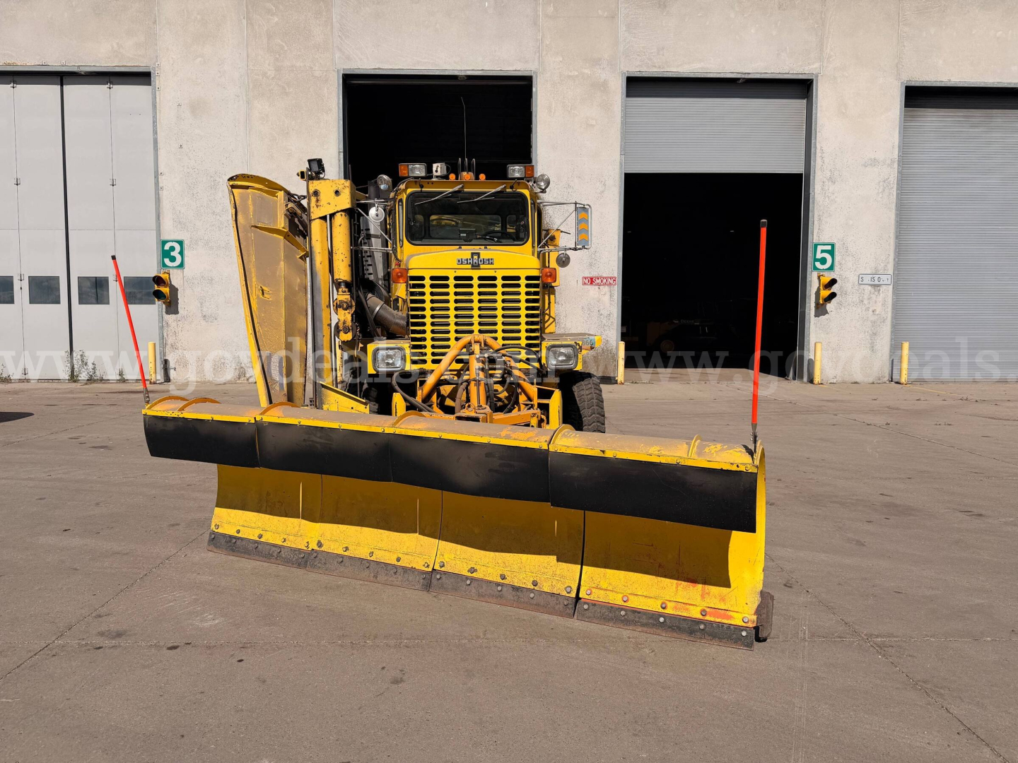 Front view of a yellow Oshkosh plow truck with a wide multi-panel snow plow attachment, warehouse bays in background.