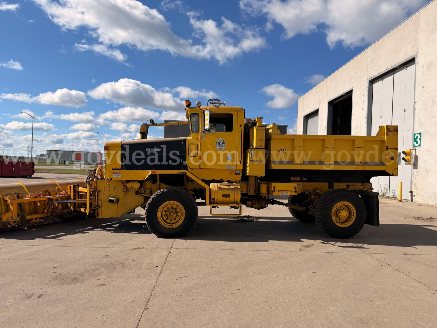 Oshkosh PA-2323-SP truck with large side-dump bed and yellow paint, side profile view, plow apparatus attached at front, clear blue sky.