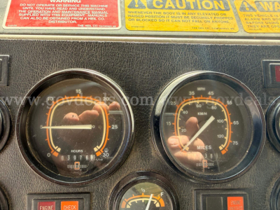 Dashboard of a truck showing two analog gauges (hours and speed) with orange accents and a small center gauge; warning labels with red and yellow text above.