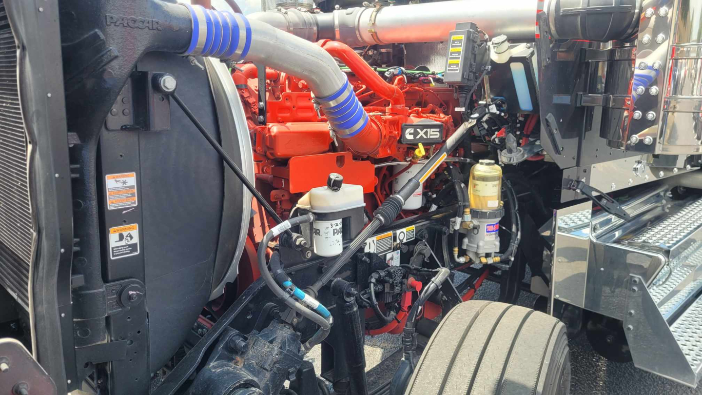 Kenworth W900L engine bay showing Xtis engine with orange/red components, hoses, reservoirs and metal framework.