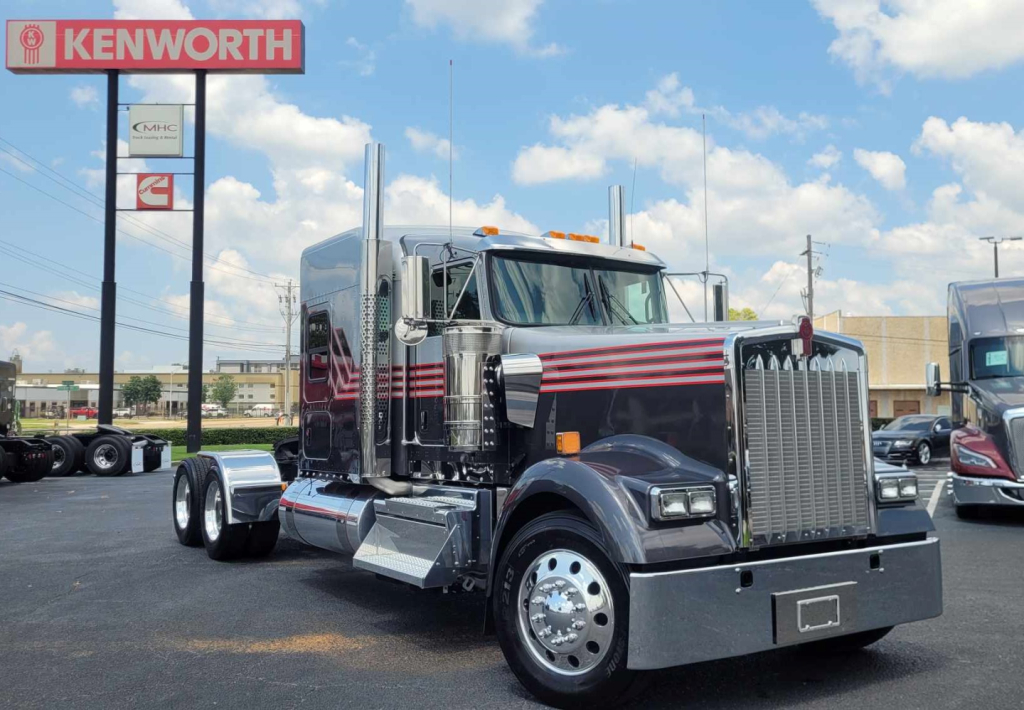 Kenworth W900L conventional sleeper truck in a dealership lot with chrome bumper and dual exhaust stacks.