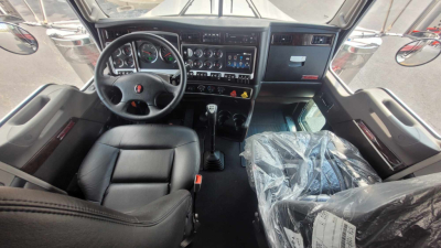 Kenworth W900L cab interior showing steering wheel, dashboard with gauges, center stack, and two seats with plastic covering on passenger seat.