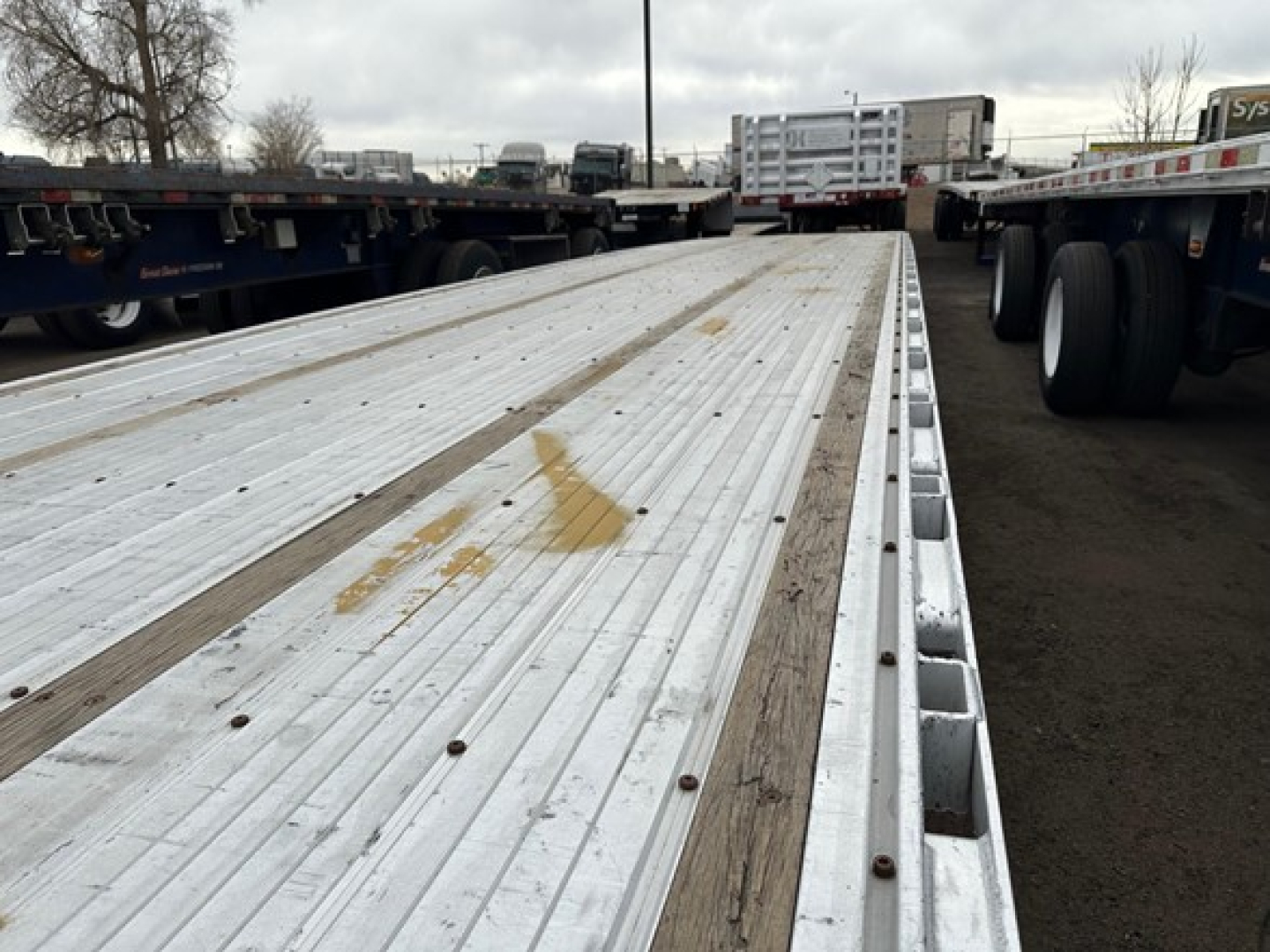 Drop deck trailer bed showing aluminum planks with wooden strips and side stakes, generic yard scene in background.