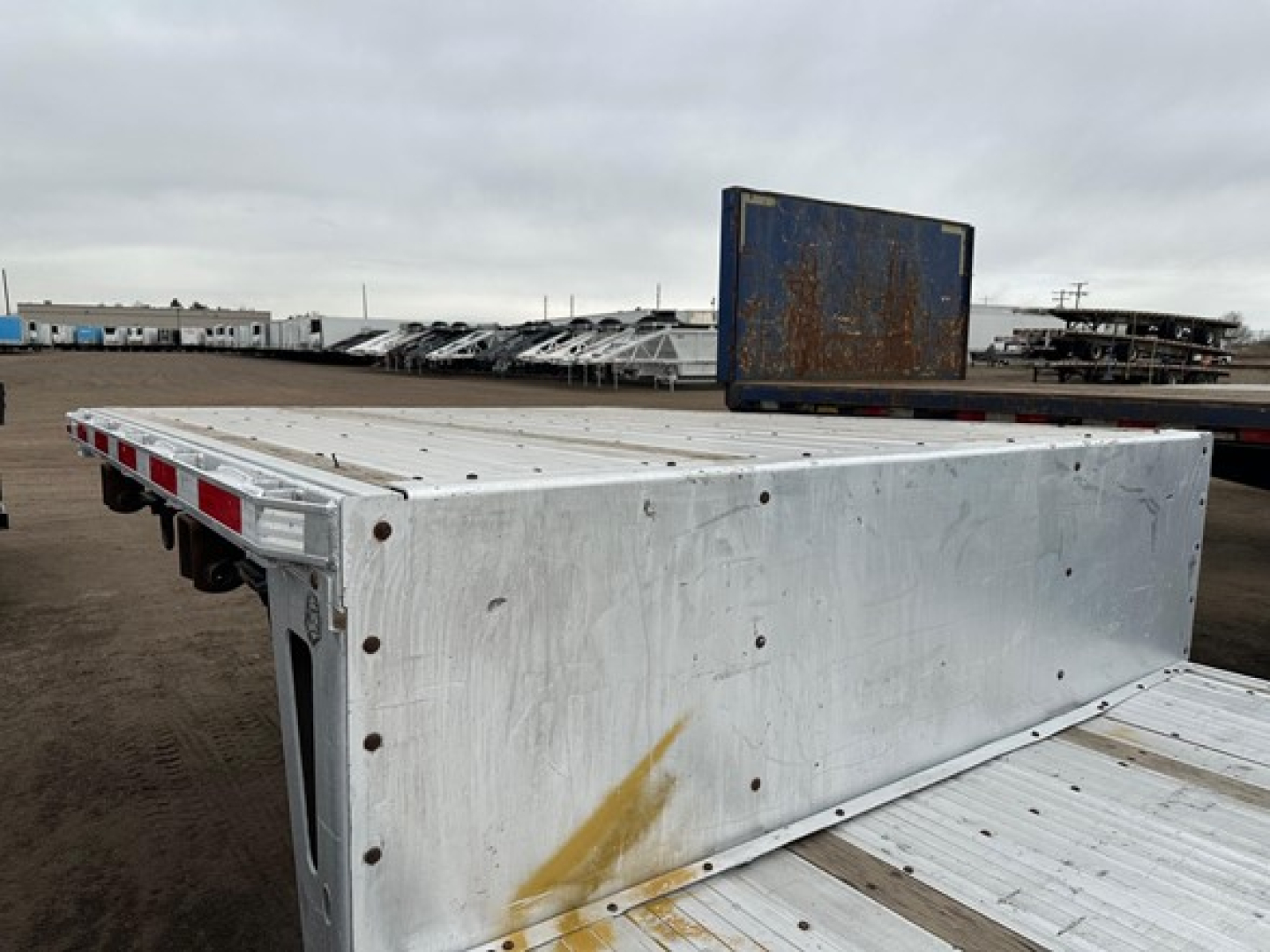 Trailer underside edge with steel drop deck trailer, aluminum side panels, and rear ramp. Visible text includes no legible branding or serial numbers in the photo.