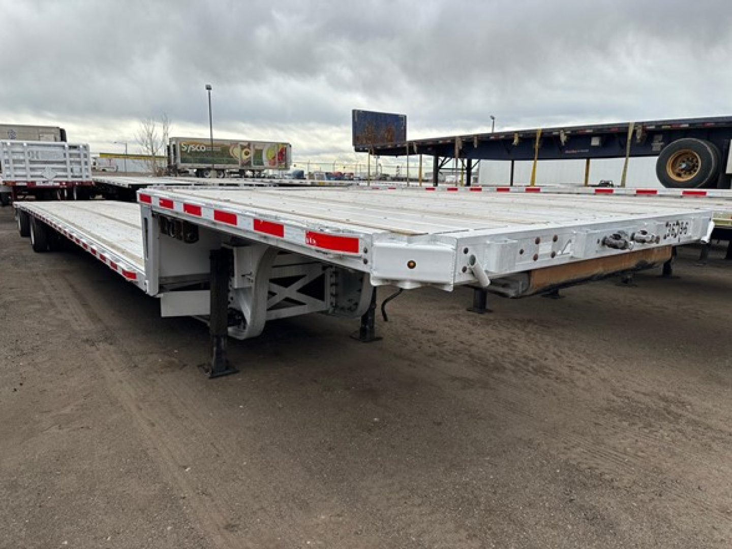Trailer: a 2015 Trail King TK Drop Deck trailer, flatbed with drop deck, showing metal frame, support legs, and reflective red/white tape along sides.