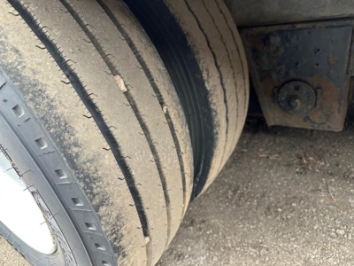 Trailer axle close-up showing dual-tire assembly with dirt on tread and sidewall; undercarriage frame and mounting brackets visible.