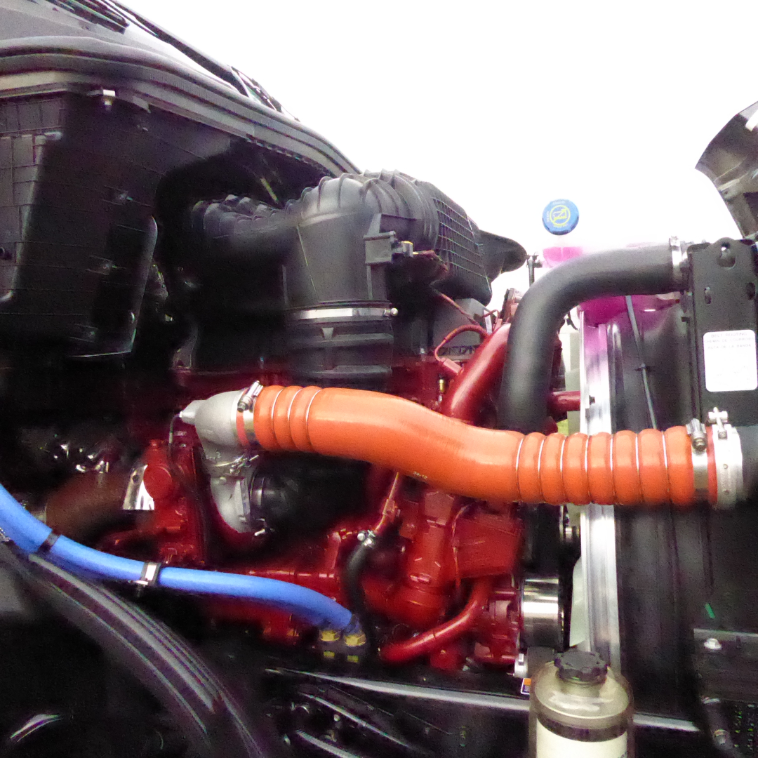 Engine bay of a 2026 Mack PR64T76H showing orange radiator hose, blue coolant line, black air intake, and red engine block.