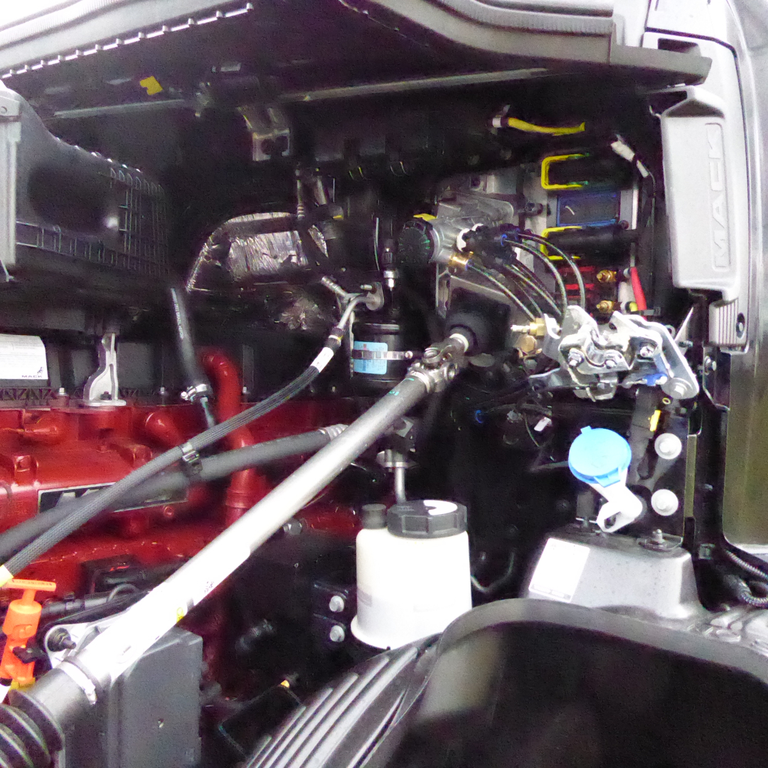 Truck engine bay showing Mack PR64T76H with visible hoses, reservoirs, and engine components.