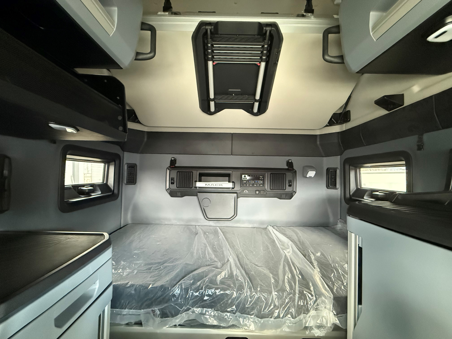 Interior of a new Mack truck sleeper cab with storage compartments and overhead shelves; center control console visible; plastic-covered floor; windows on sides.