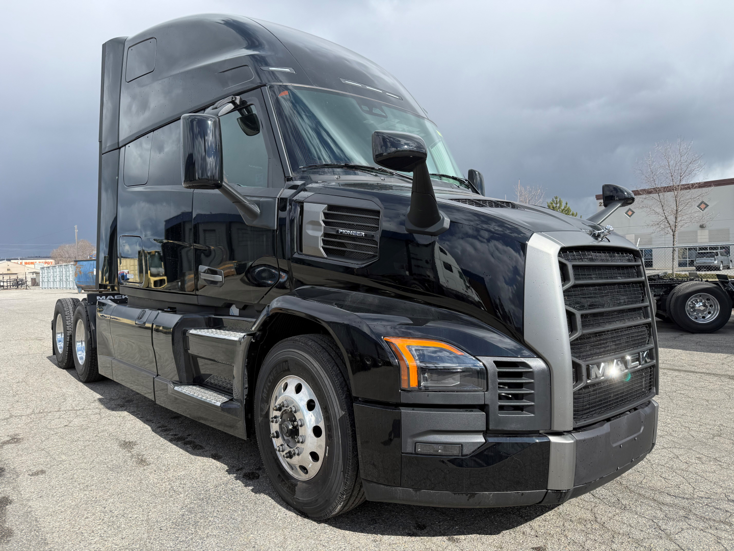 Truck cabin and front view of a black 2026 Mack PR64T76H sleeper tractor showing chrome grille, side mirrors, and wheel rims.