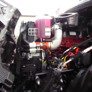 Engine bay of a new Mack PR64T76H truck showing pink air intake filter, turbo piping, hoses, and surrounding components.