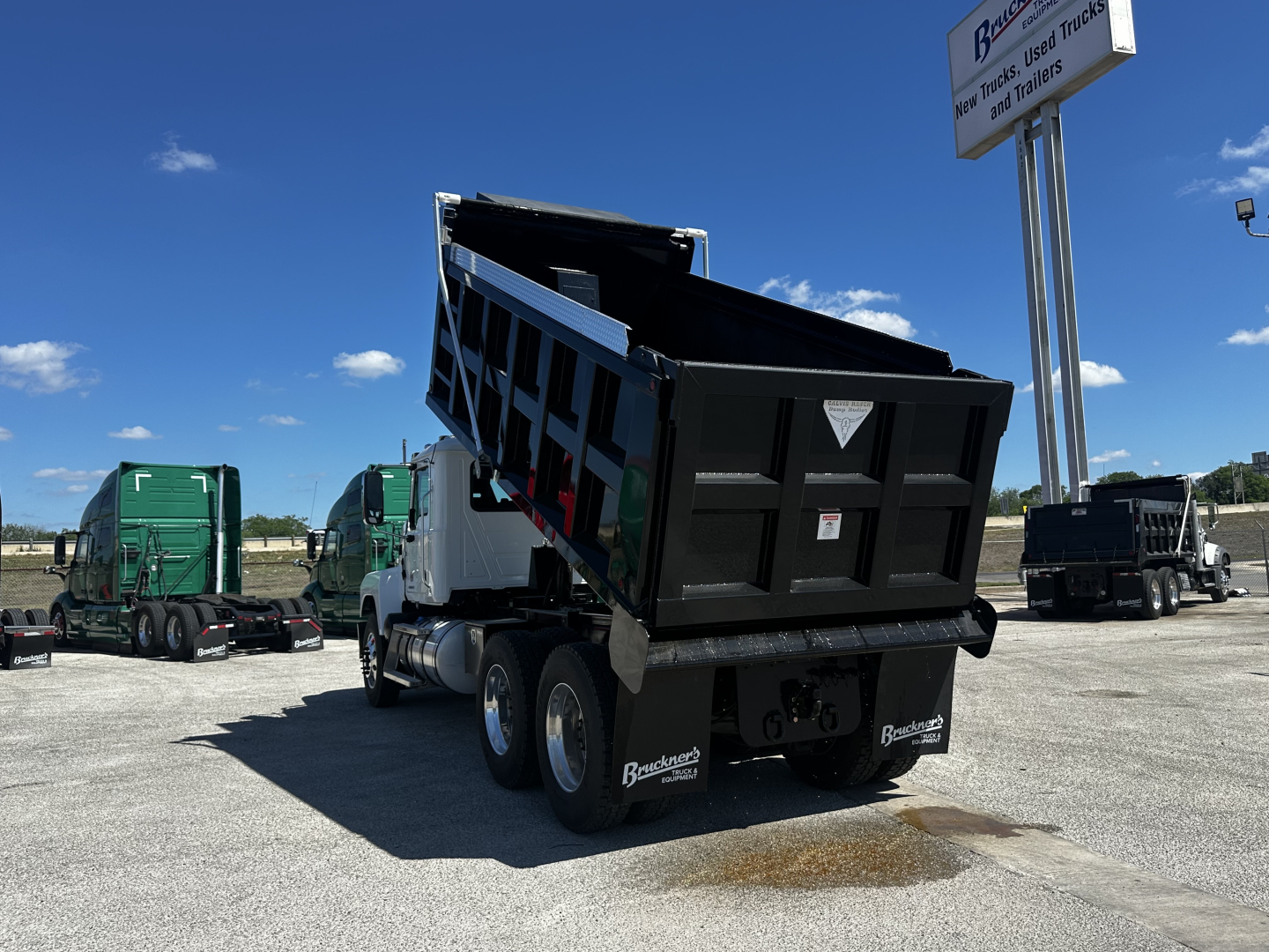 Truck dump body on Mack PI64 chassis with raised hydraulic bed, rear view showing dual rear axles and stake bed, Bracken trucks lot in background.