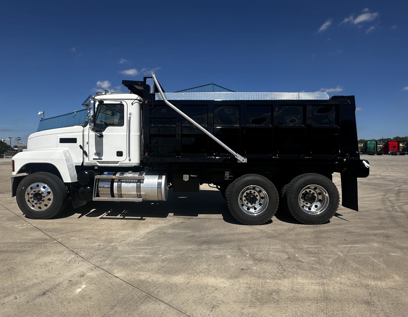 Mack PI64 dump truck side view showing black dump body, white cab, chrome fuel tanks, dual rear axles, and diamond plate toolbox.