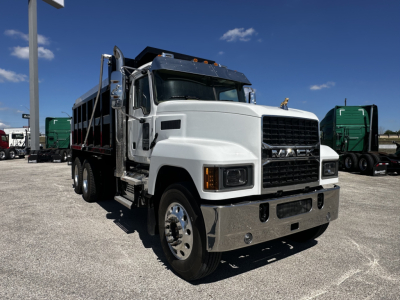 Mack PI64 dump truck in a showroom lot showing front grille, large bumper, black grille insert, dual rear wheels, and cargo bed.