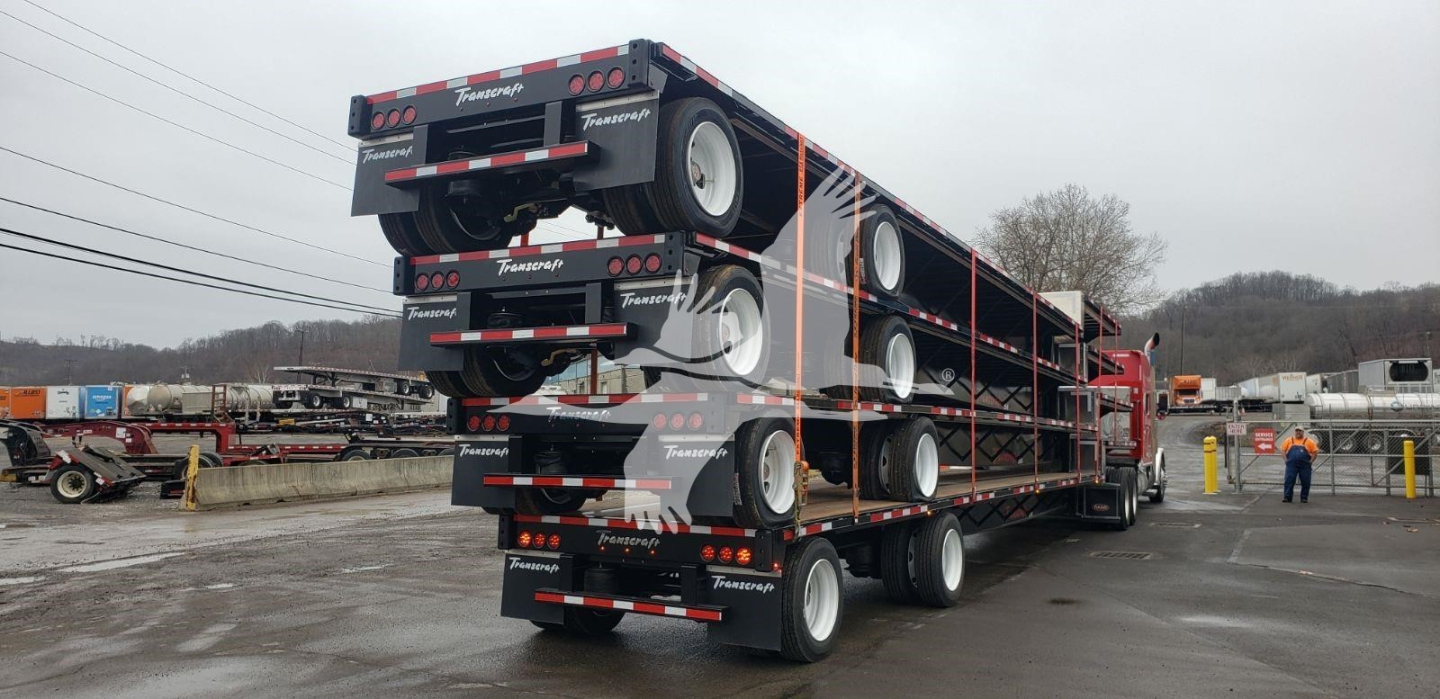 Transcraft DTL-2100 drop deck trailer stacked on its side in a yard, showing multiple axles and dual rear ramps; text 'Transcraft' branding on the sides and tires visible.