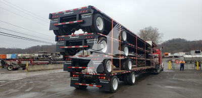 Transcraft DTL-2100 drop deck trailer stacked on its side in a yard, showing multiple axles and dual rear ramps; text 'Transcraft' branding on the sides and tires visible.