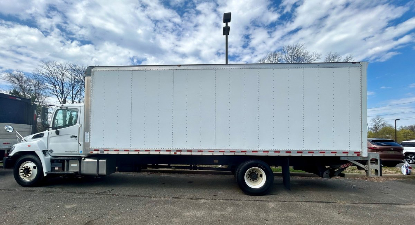 Box truck side view showing 2020 Hino 338 box van with white cargo area and cab, empty rear wheel area, clear sky.
