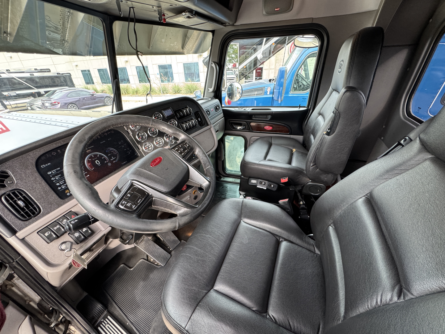 Peterbilt 389 cab interior showing steering wheel gauges and driver's seat with control panel and view through side window.
