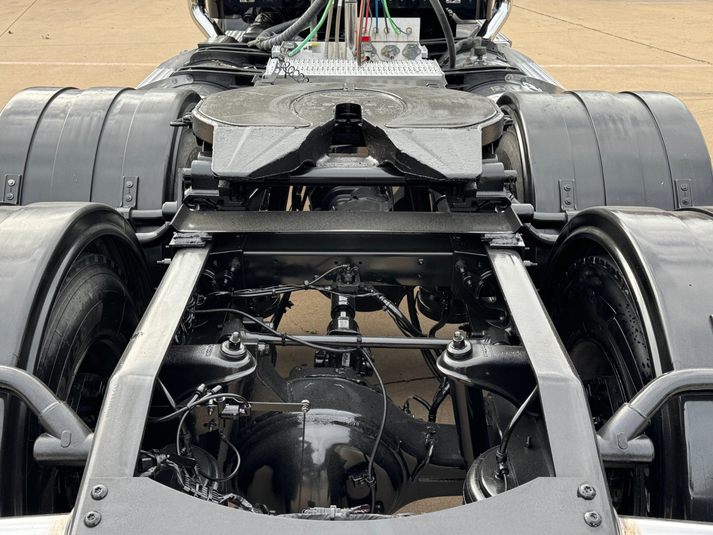 Peterbilt 389 chassis and drivetrain view showing rear axle assemblies and fuel/air lines; exposed suspension and wiring harnesses in a truck yard.