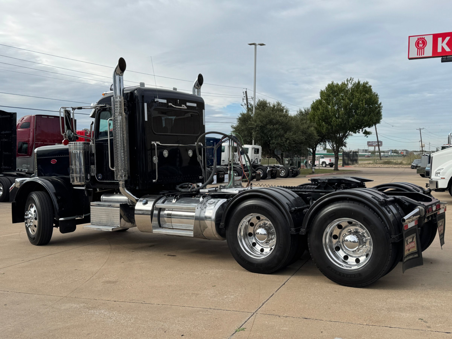Peterbilt 389 conventional daycab tractor in a used truck lot, low-angle side view showing dual rear axles and chrome exhaust stacks.