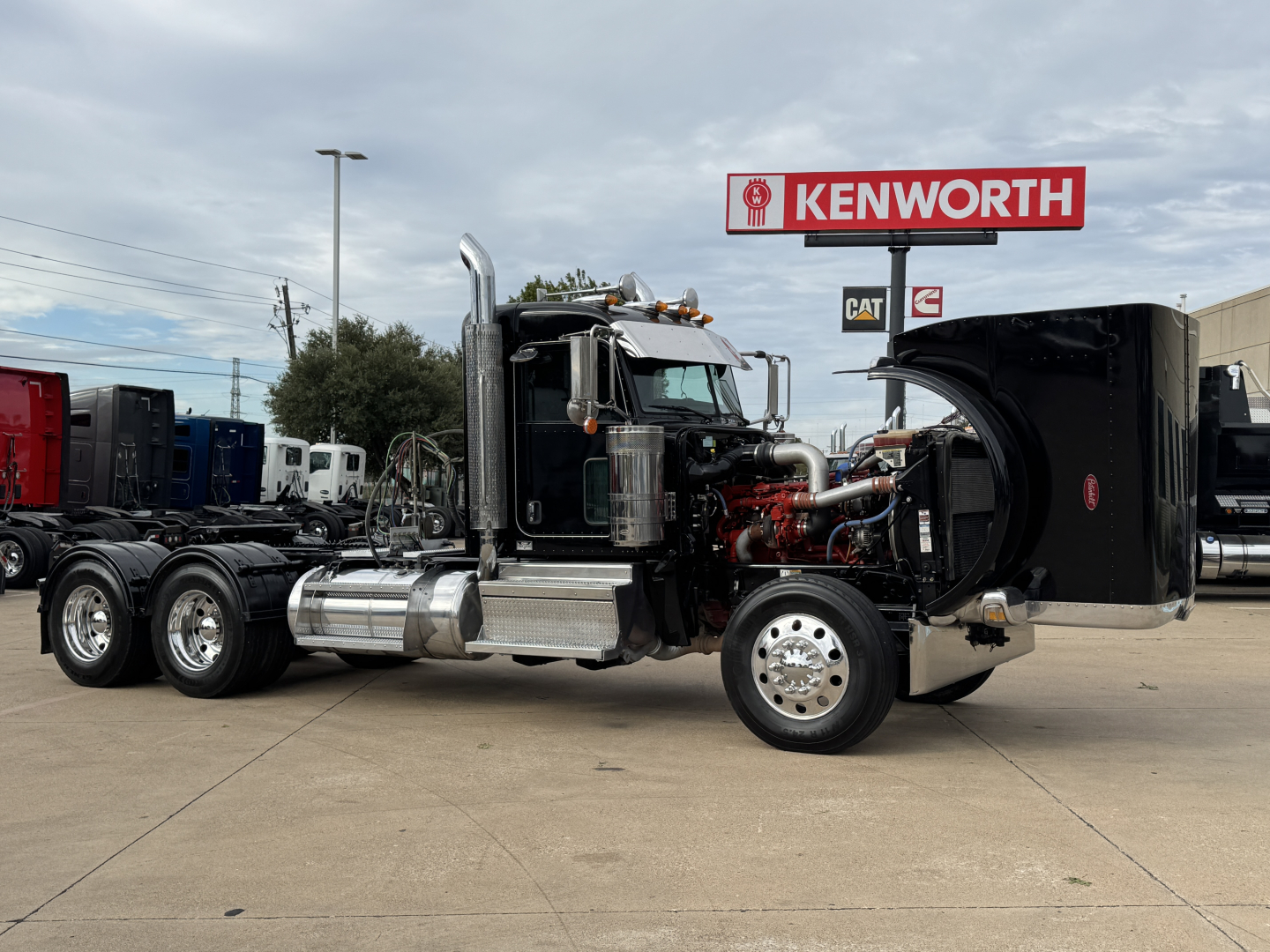 Peterbilt 389 conventional daycab truck in a dealership lot with hood open showing engine exposure and chrome exhaust stacks.