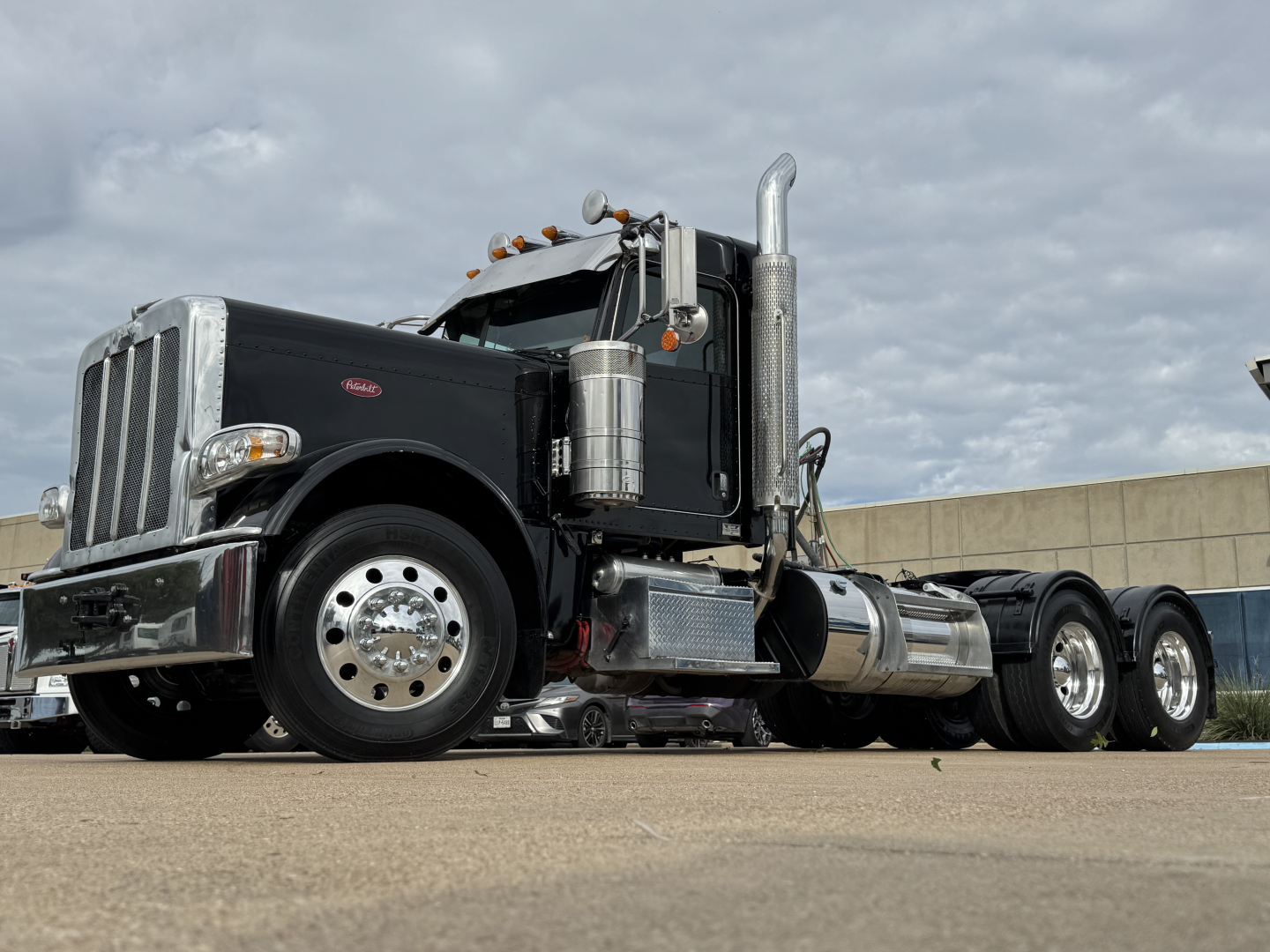 Peterbilt 389 conventional daycab truck in black, front-left view, fuel tanks, chrome exhaust stacks, dual rear axles, clear sky.