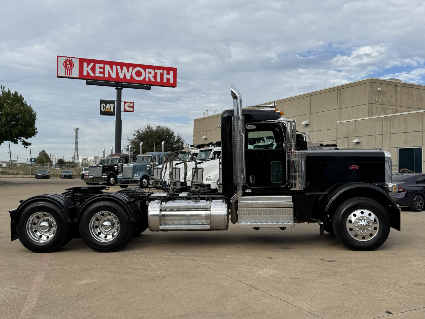 Peterbilt 389 conventional daycab truck side view showing dual rear axles and chrome fuel tanks under a Kenworth sign.