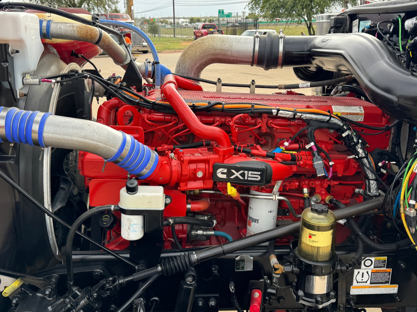 Peterbilt 389 engine bay showing Cummins X15 engine with red intake manifold, blue turbo hoses, and various components including filters and hydraulic lines.