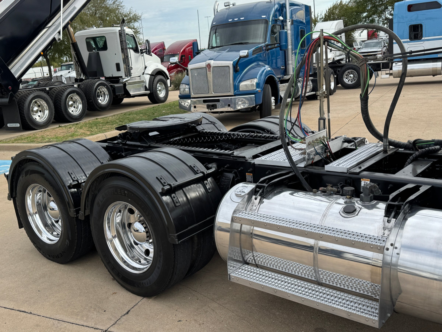 Peterbilt 389 truck chassis and fuel tank showing rear tandem axles and chrome fuel tank; other trucks in background at dealer lot.