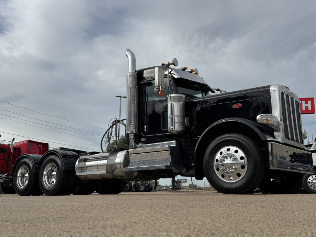 Peterbilt 389 conventional daycab truck with chrome fuel tanks and dual rear axles, front left view, black paint, chrome accents, daytime cloudy sky.