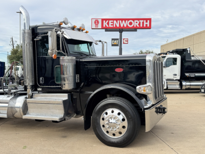 Peterbilt 389 conventional daycab in black, front-left view showing chrome exhaust stacks, large chrome bumper, Dual round headlights, polished wheels, Kenworth sign in background.