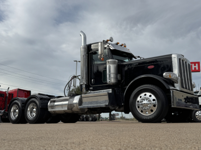 Peterbilt 389 conventional daycab truck with chrome fuel tanks and dual rear axles, front left view, black paint, chrome accents, daytime cloudy sky.