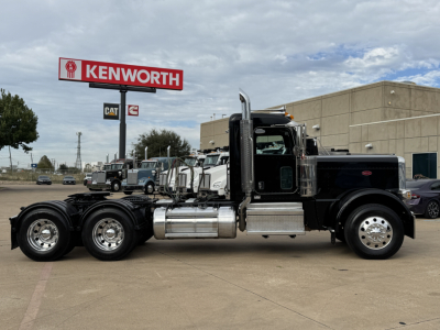 Peterbilt 389 conventional daycab truck side view showing dual rear axles and chrome fuel tanks under a Kenworth sign.