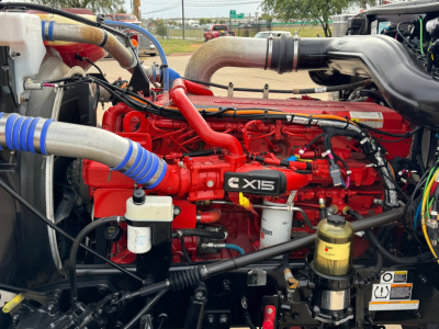 Peterbilt 389 engine bay showing Cummins X15 engine with red intake manifold, blue turbo hoses, and various components including filters and hydraulic lines.