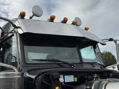 Peterbilt 389 cab front view showing sun visor, multiple roof cab lights, and windshield with wipers. Visible stickers on dash area and hood overlap.